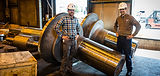 Two men stand with large metal parts in a workshop, Scot Forge.