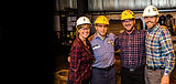 Four Scot Forge employees stand together, smiling, wearing hard hats. Workplace environment.