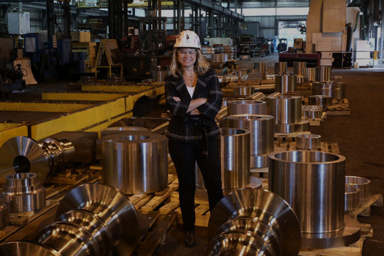 Woman wearing helmet stands among metal components in factory Scot Forge